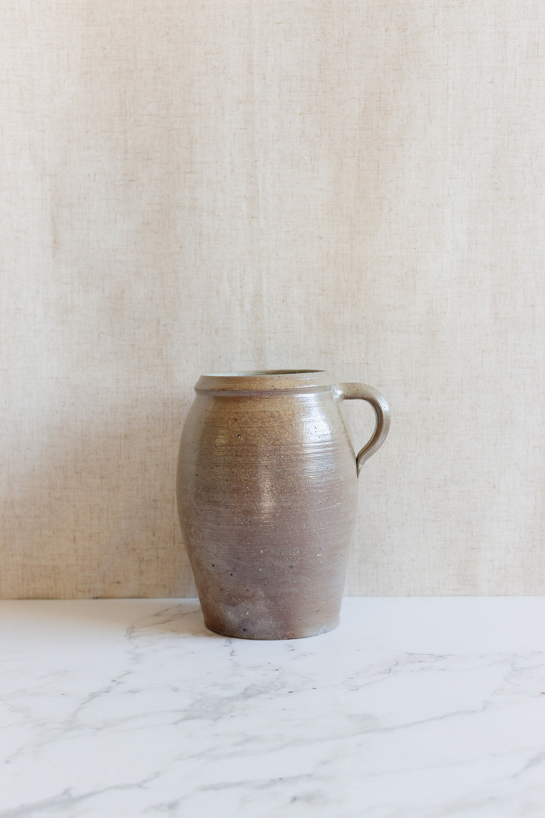 A midcentury French stoneware vessel by H. Chevalier & Fils, Orléans rests on a white marble surface with a beige textured background, highlighting its rounded shape, narrow opening, and natural earthy glaze.