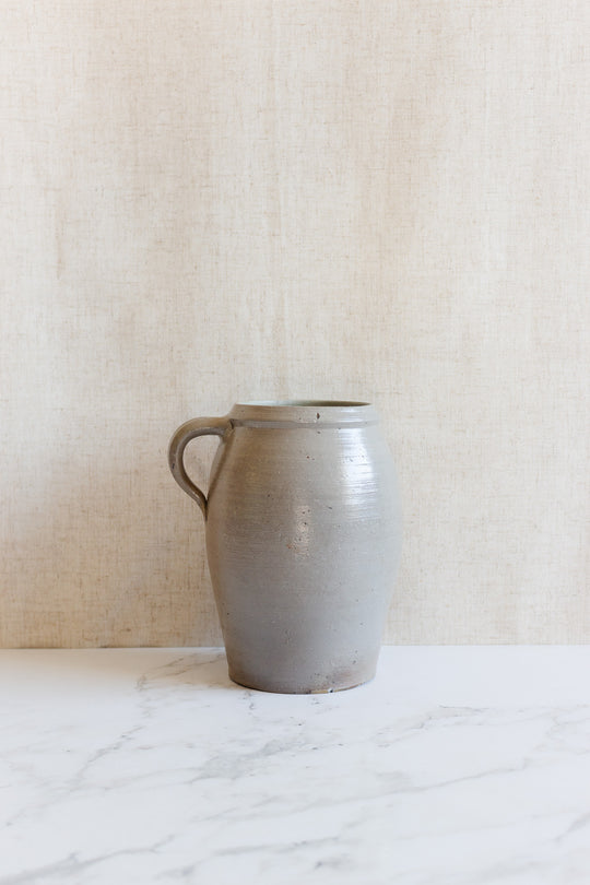 The midcentury French stoneware vessel by H. Chevalier & fils, Orléans, in simple gray with a handle, rests on a white marble surface before a beige textured background.