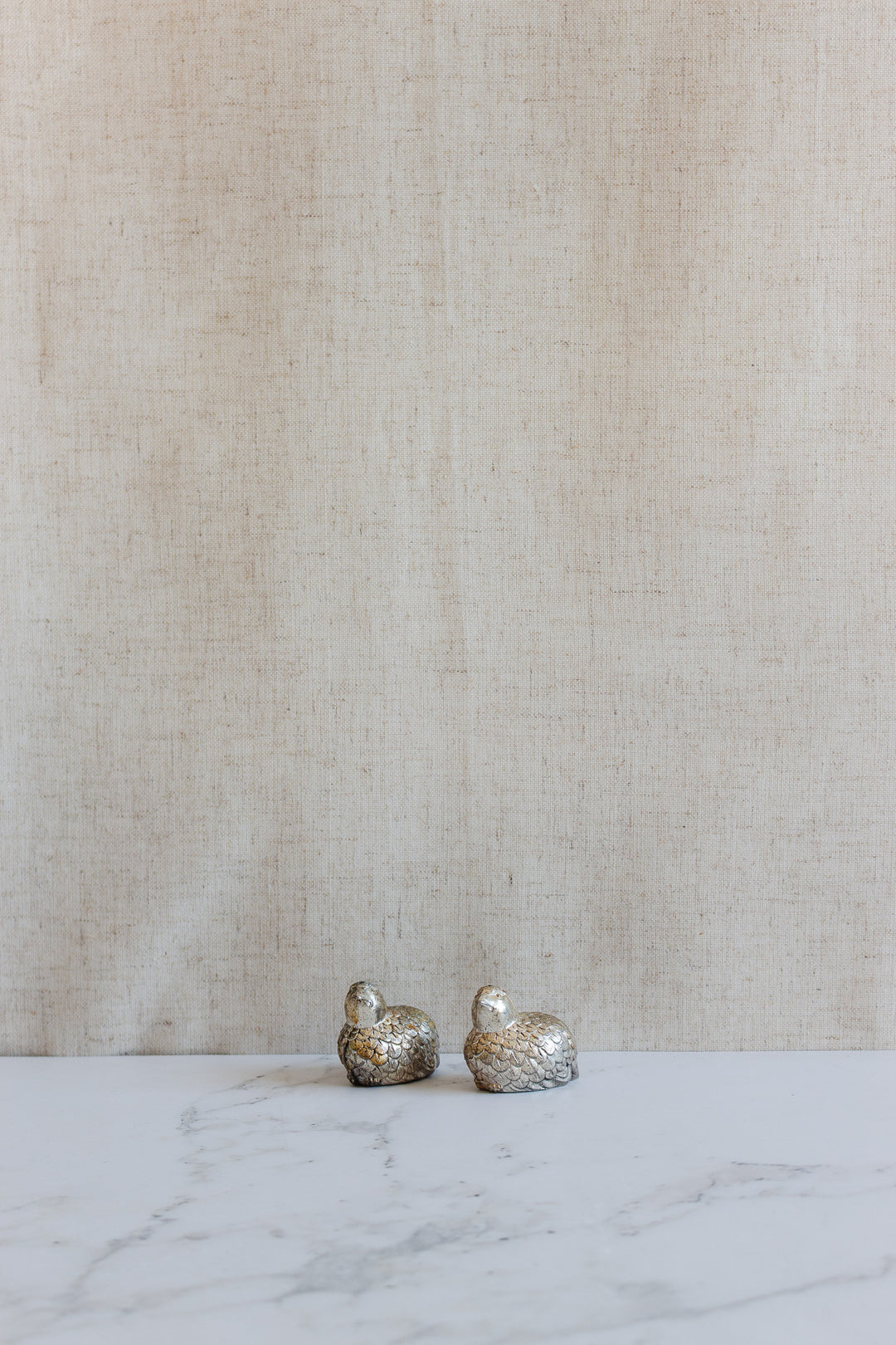 Two 1970s Italian silver plate quail salt and pepper shakers are displayed on a white marble surface with a beige textured fabric background.
