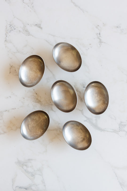 Six vintage French stainless steel oblong ice cream cups, featuring a slightly tarnished matte finish, are arranged in a loose circular pattern on a white marble surface.