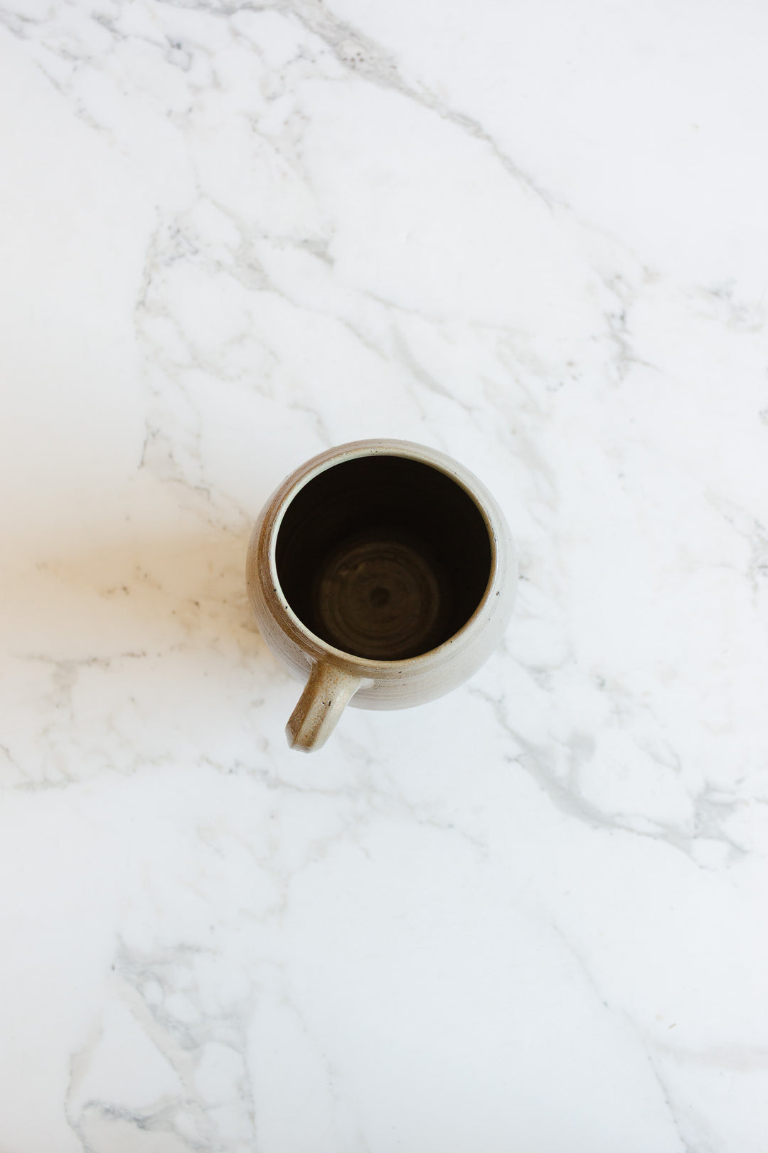 A midcentury French stoneware vessel by H. Chevalier & fils, Orléans with a handle sits empty on a white marble surface, viewed from above; the background features subtle gray veining characteristic of marble.