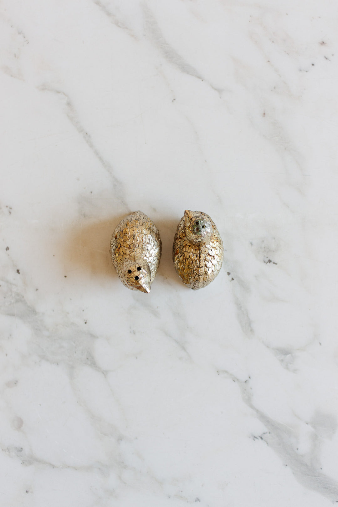 Two small, silver-plated 1970s Italian quail salt and pepper shakers sit side by side on a white marble surface.
