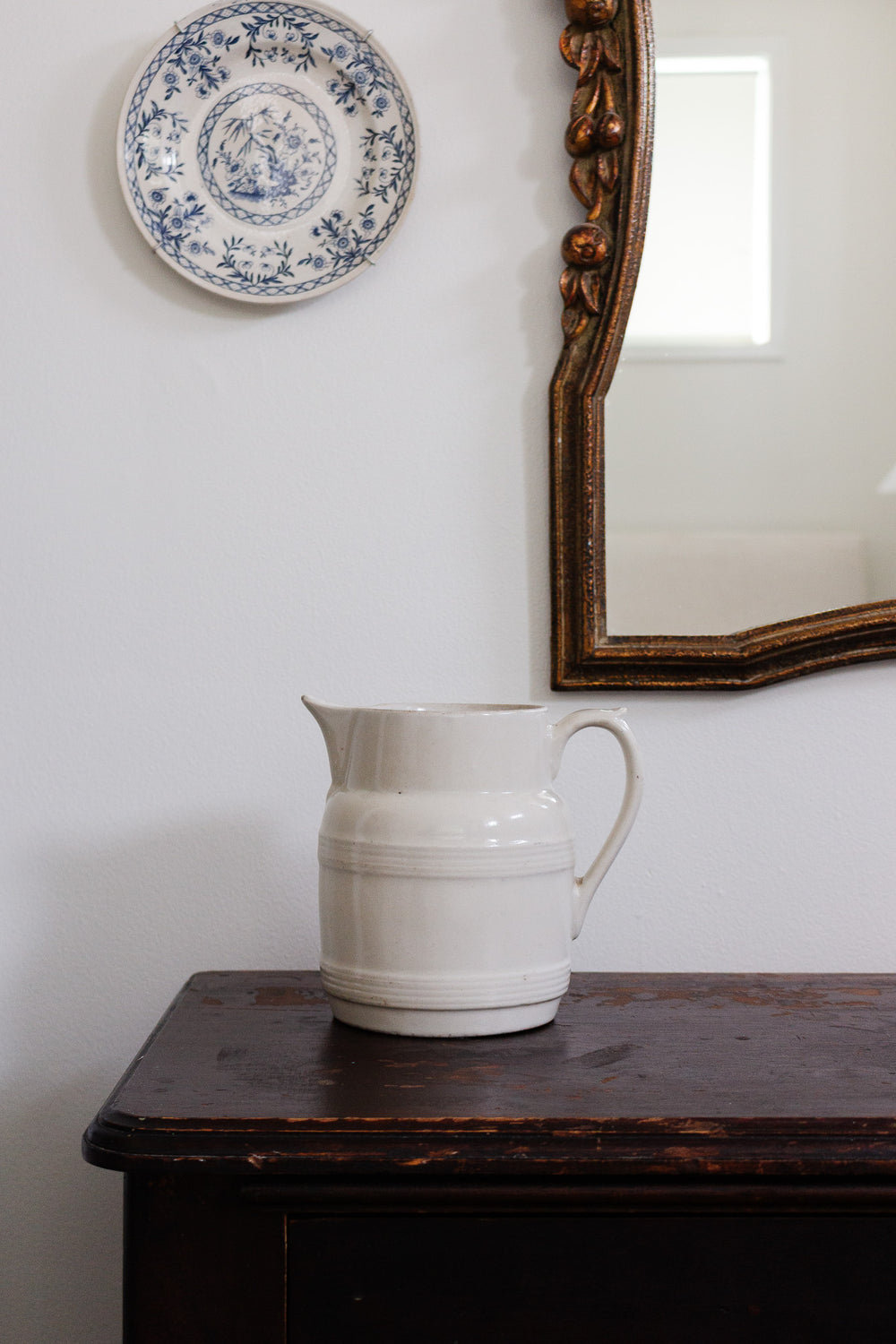 An antique French white stoneware pitcher rests on a dark wooden table beneath a blue and white decorative plate on the wall and a gold-framed mirror reflecting a window.