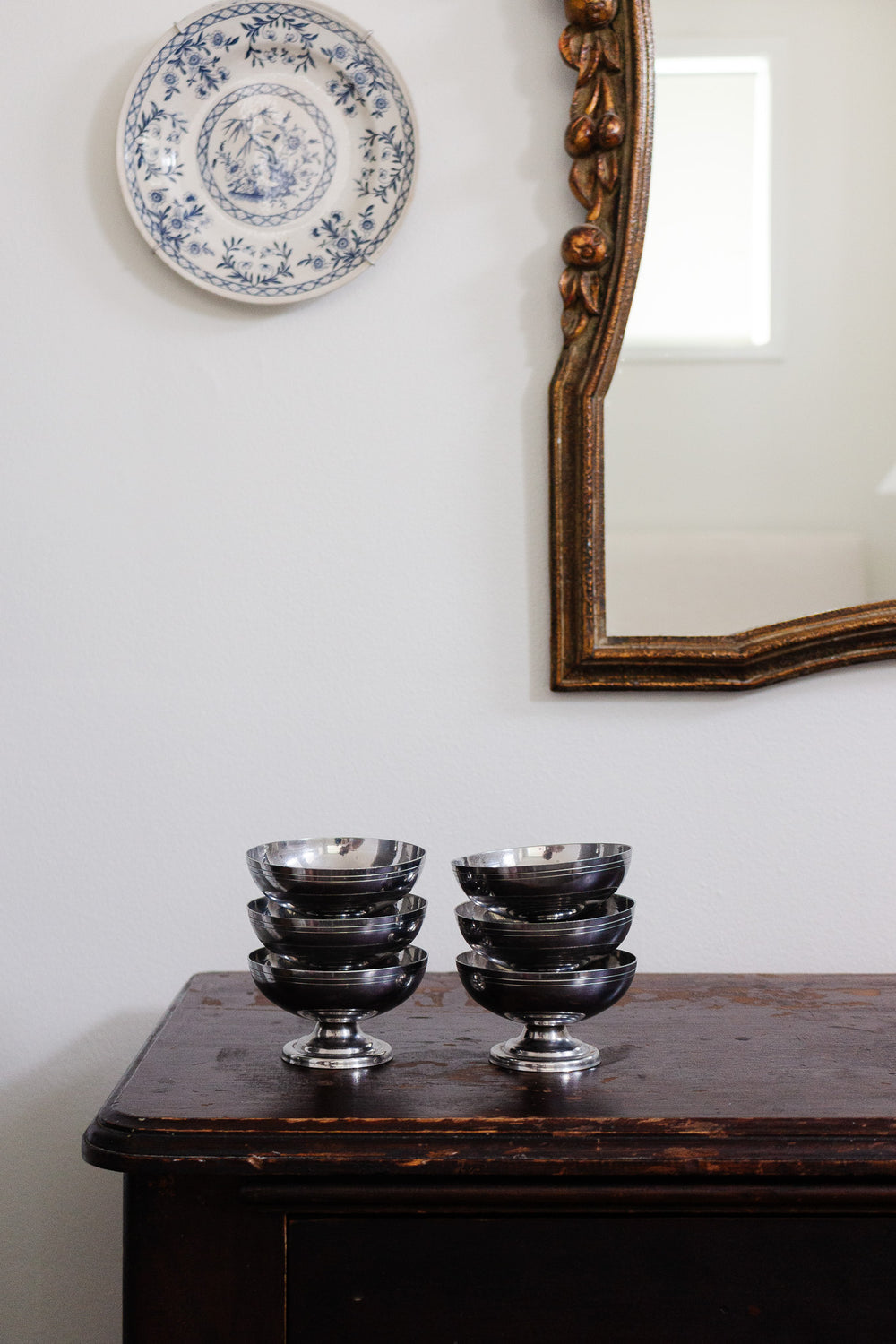 A set of 6 midcentury French ice cream cups is displayed on a dark wooden table below a decorative blue and white plate and a gold-framed mirror on a white wall.