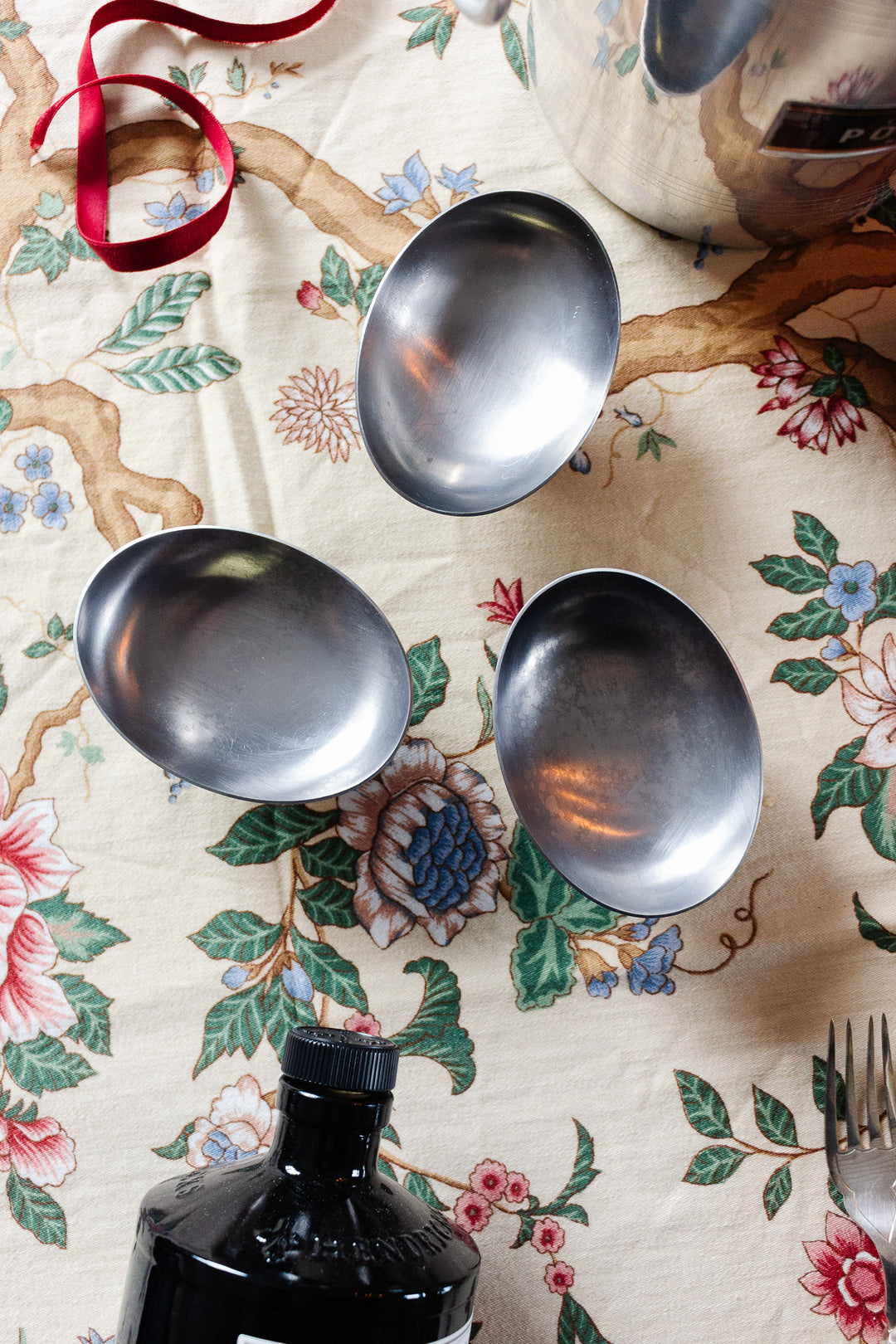 Three vintage French stainless steel oblong ice cream cups sit on a floral-patterned tablecloth, along with a red ribbon, a black bottle, a fork, and part of a reflective metal pot.