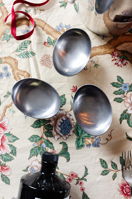 Three vintage French stainless steel oblong ice cream cups sit on a floral-patterned tablecloth, along with a red ribbon, a black bottle, a fork, and part of a reflective metal pot.