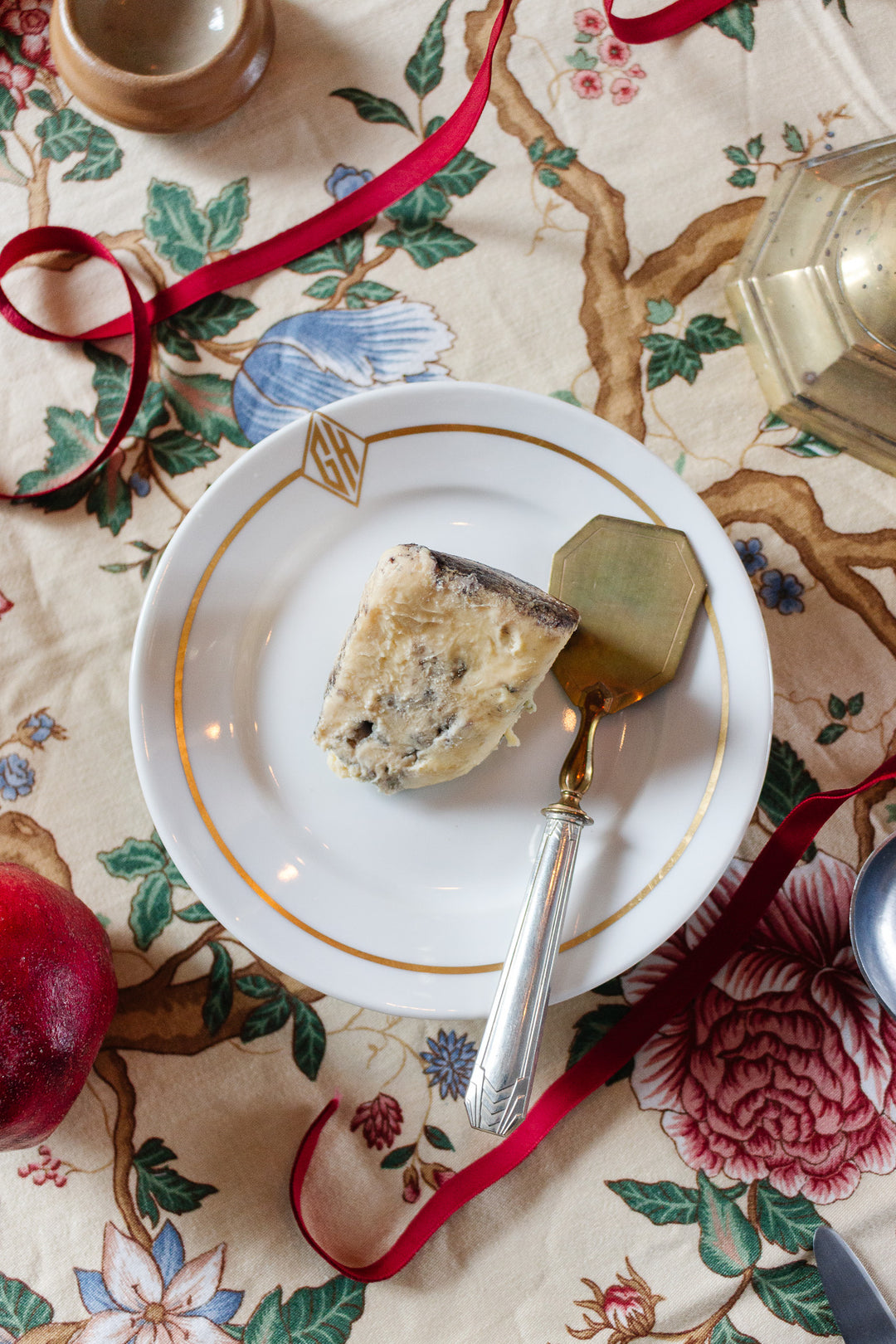 A slice of blue cheese rests on a white plate from a 19th century French gold vermeil & silver dessert set, with a cheese knife atop a floral tablecloth, surrounded by a red ribbon, pomegranate, and glass.