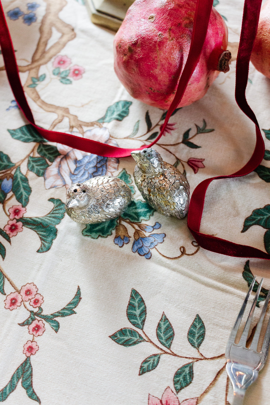 A patterned tablecloth with a red velvet ribbon, 1970s Italian silver plate quail salt and pepper shakers, a pomegranate, and a fork evoke classic 1970s table decor.