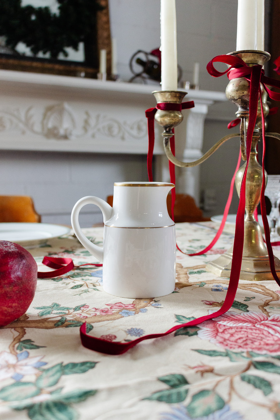 A white mug with a gold rim sits on a floral tablecloth beside a vintage Wedgwood “Metallised” bone china milk jug, a brass candelabra with red ribbons, a red pomegranate, and a white fireplace in the background.