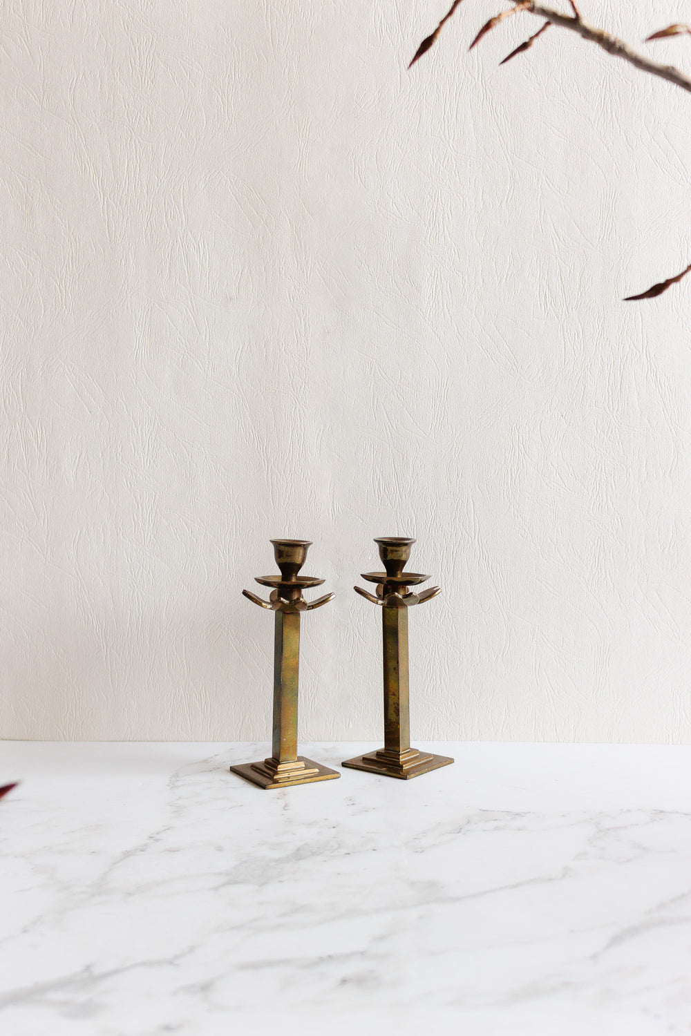 A pair of candleholders with angular designs sit side by side on a white marble surface, their elegant forms standing out against a textured beige wall while thin reddish branches frame the scene.