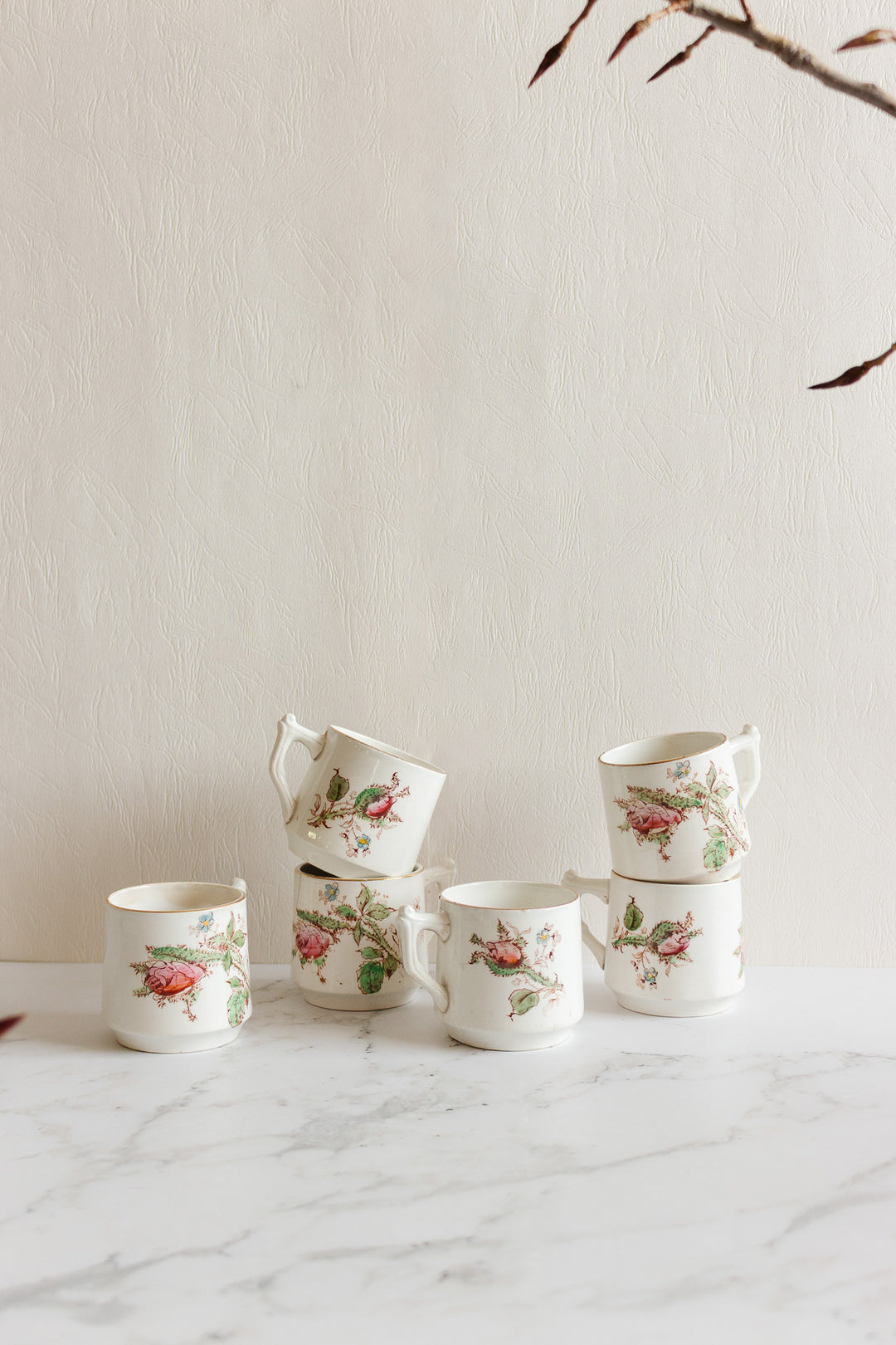 Five antique Gien transferware coffee cups, featuring delicate floral and bird motifs, are stacked on a white marble surface against a light textured wall, with dried branches framing the upper right corner of the image.