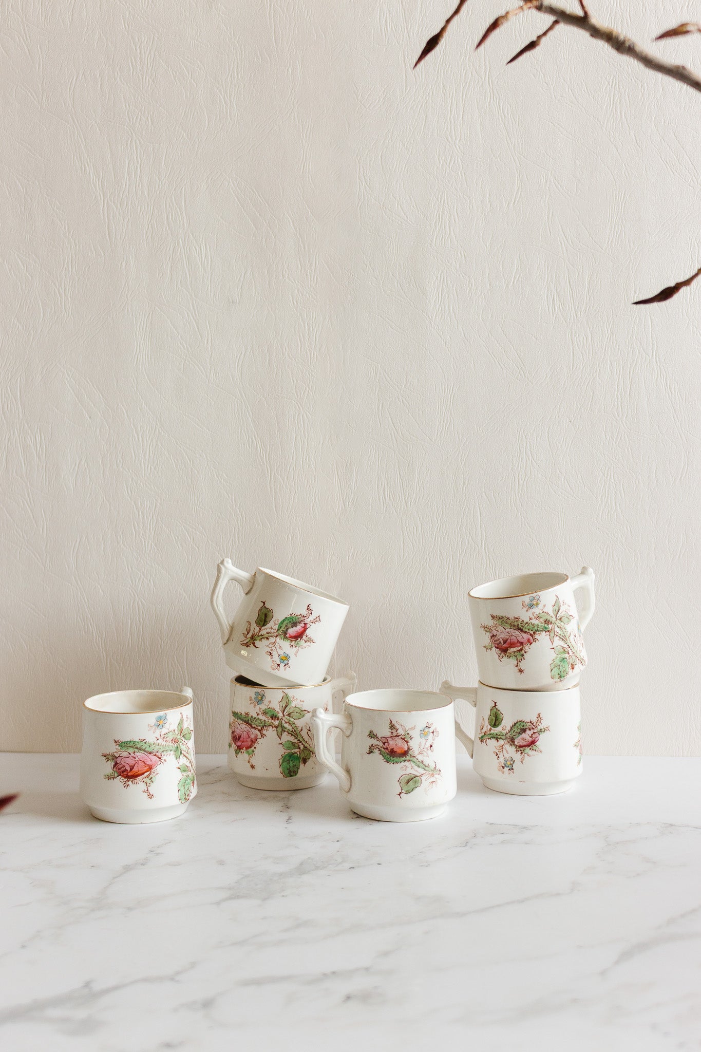 Five antique Gien transferware coffee cups, featuring delicate floral and bird motifs, are stacked on a white marble surface against a light textured wall, with dried branches framing the upper right corner of the image.