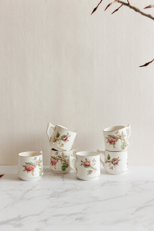 Five antique Gien transferware coffee cups, featuring delicate floral and bird motifs, are stacked on a white marble surface against a light textured wall, with dried branches framing the upper right corner of the image.
