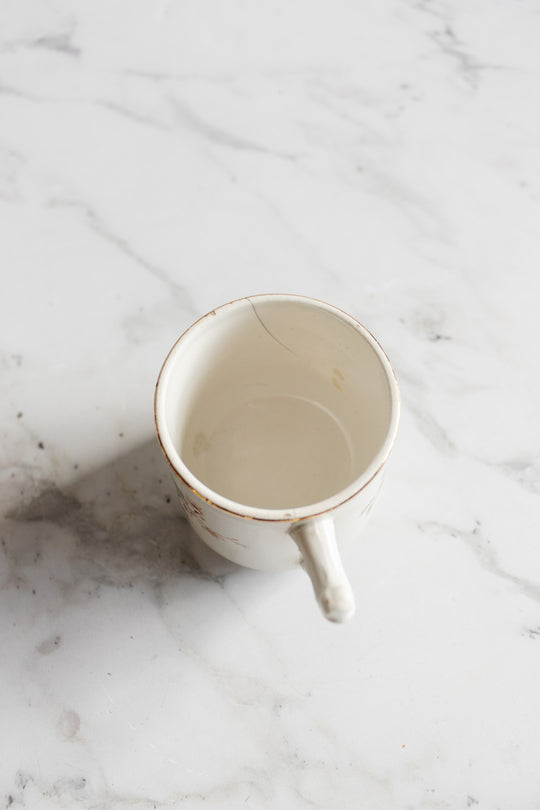An antique Gien transferware coffee cup with a visible crack on its rim sits on a white marble surface, viewed from above to showcase its dimensions.