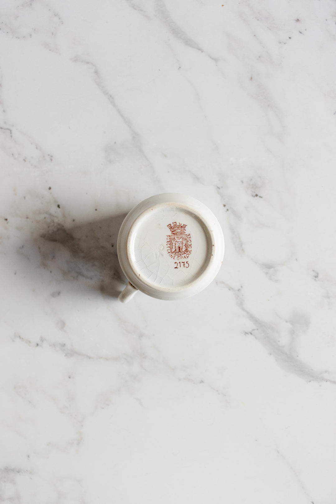An antique gien transferware coffee cup is turned upside down on a marble surface, displaying a red crest and the number 2115 on its base, with elegantly proportioned dimensions.