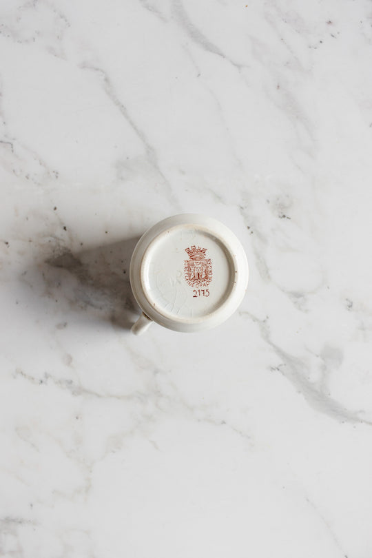 An antique gien transferware coffee cup is turned upside down on a marble surface, displaying a red crest and the number 2115 on its base, with elegantly proportioned dimensions.