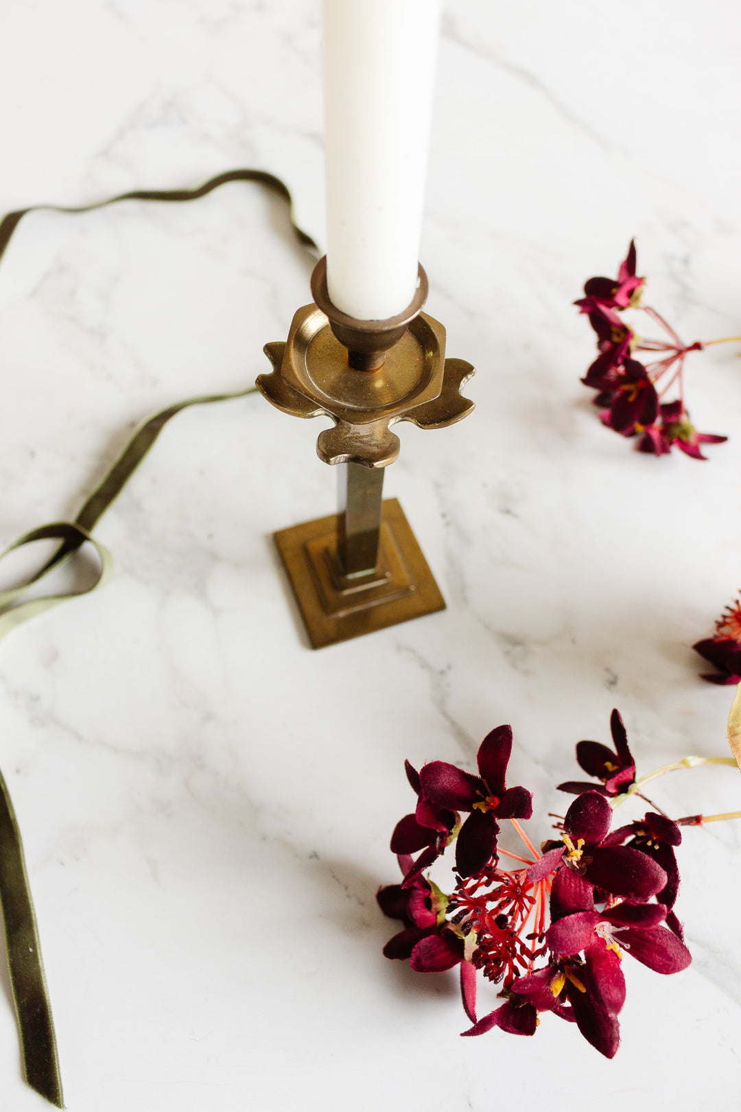 A pair of candleholders with white candles sits on a marble surface, surrounded by deep red flowers and a dark green ribbon, creating a striking and elegant arrangement.