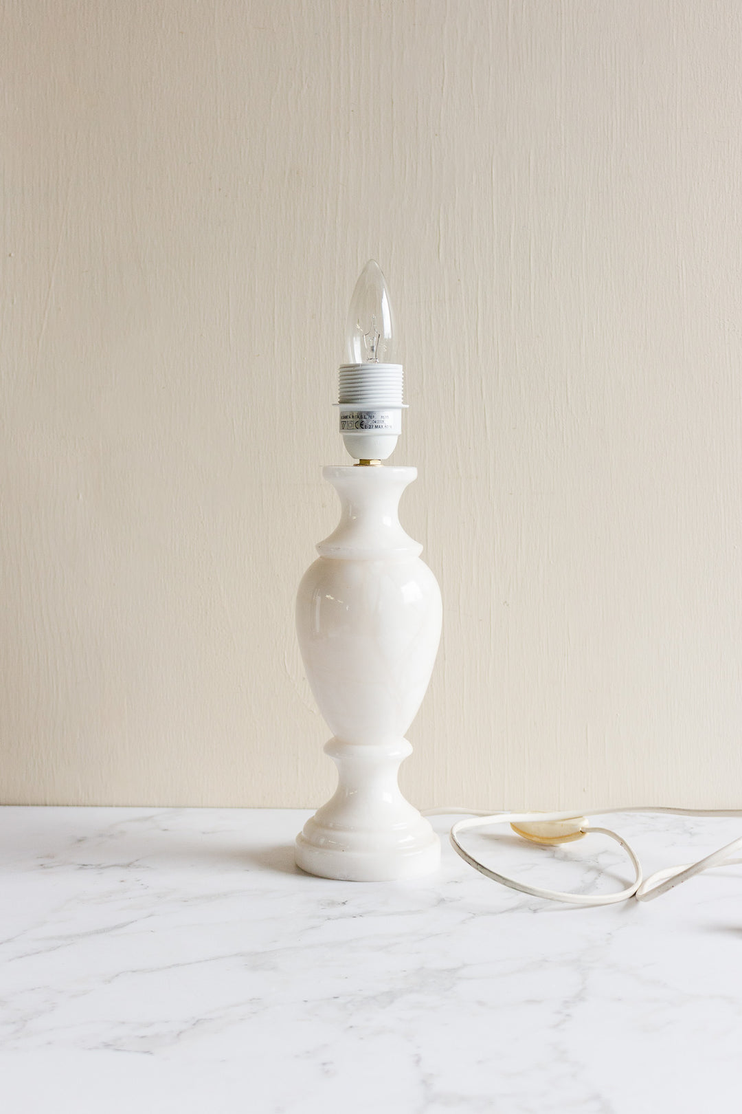 A vintage French alabaster table lamp with a bulb socket but no lampshade sits on a white marble surface against a light beige wall.