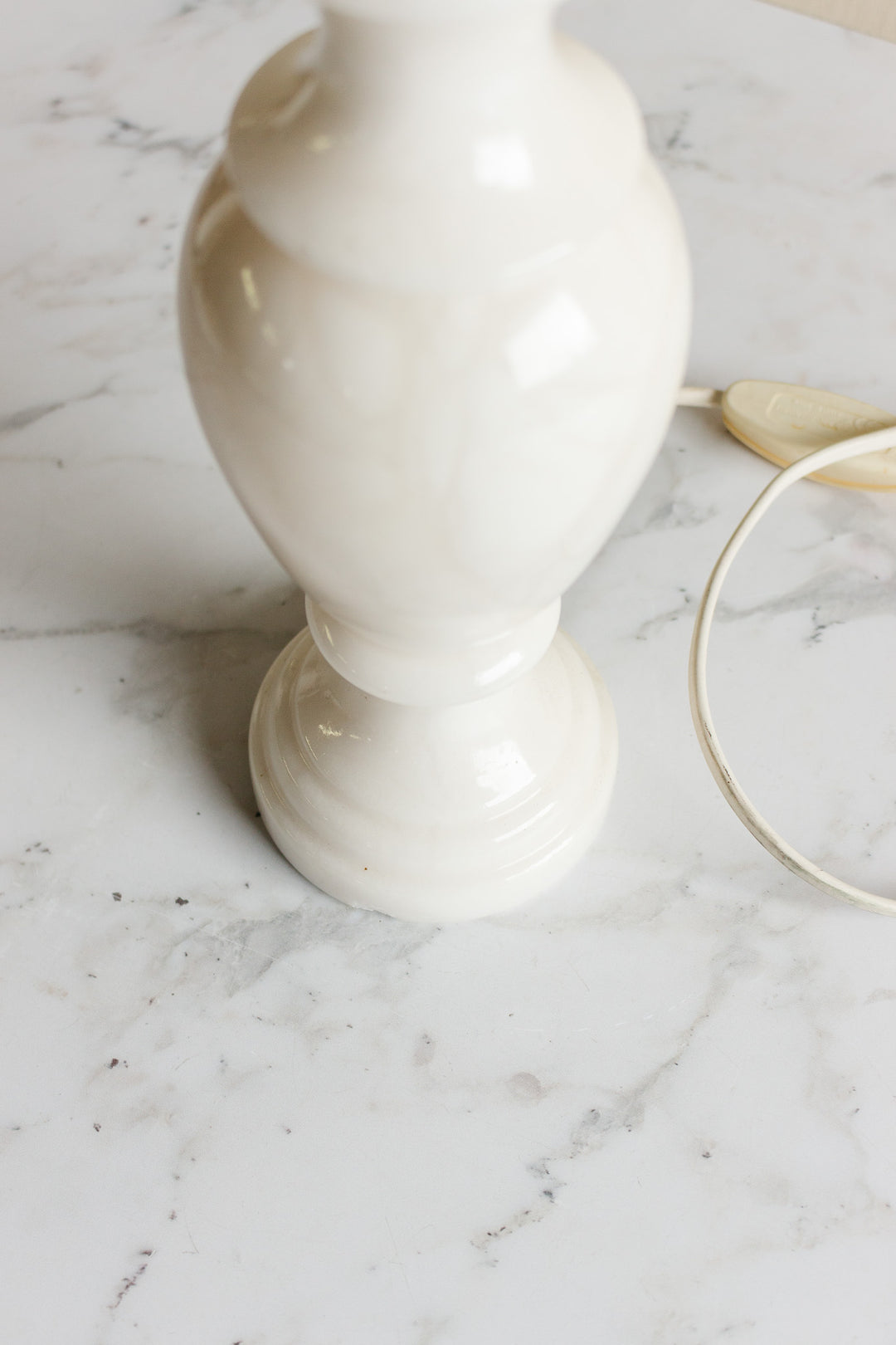 A close-up of the base of a vintage French alabaster table lamp on a white marble surface, with its electric cord and switch visible on the right.