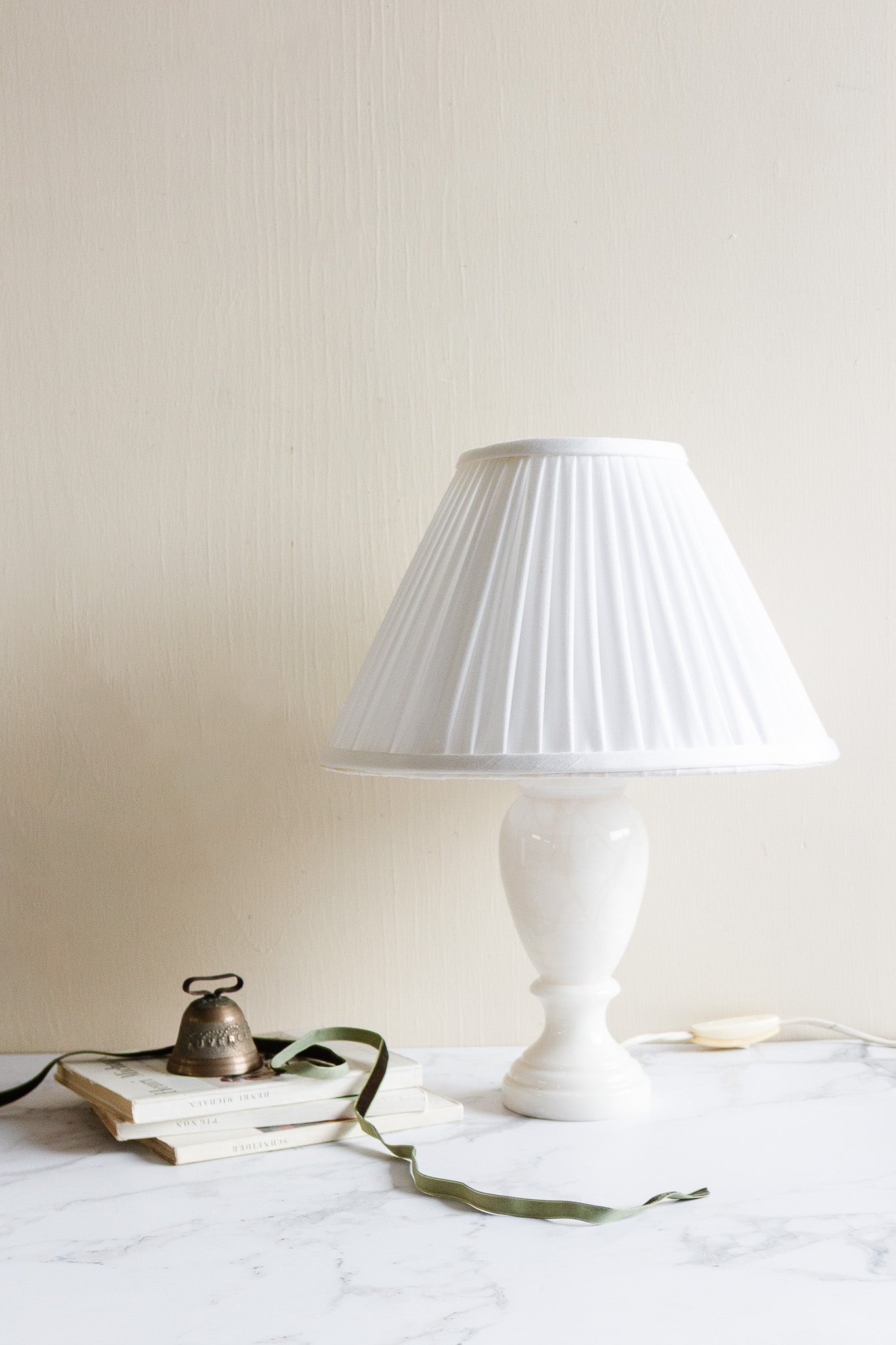 A vintage French alabaster table lamp with a pleated shade sits on a marble surface beside stacked books, a small bell, and a green ribbon against a plain light wall.