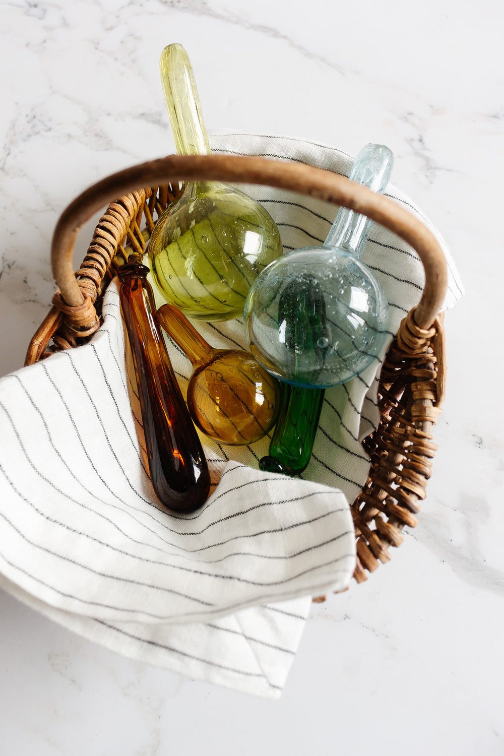 A wicker basket lined with a white cloth featuring black stripes holds four vintage French blown glass "courge" watering globes in yellow, green, blue, and amber, adding rustic elegance and Provence style to a white marble surface.