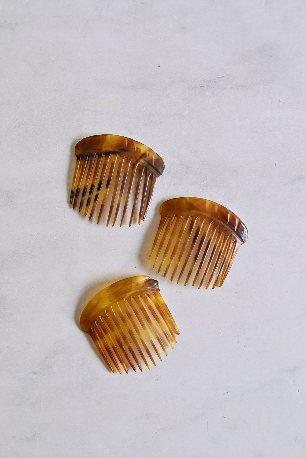 Three "turn of the century French horn hair combs" in tortoise shell, featuring wide teeth, are arranged on a light marble-like surface for a classic hair accessory look.