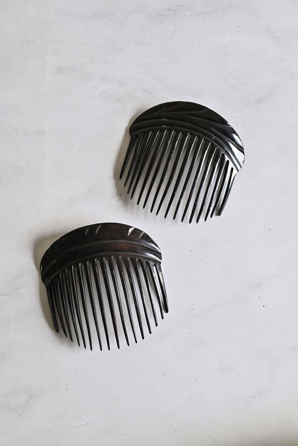 Two black "turn of the century french horn hair combs" with curved tops and evenly spaced teeth are placed slightly apart and angled on a light gray, textured surface.